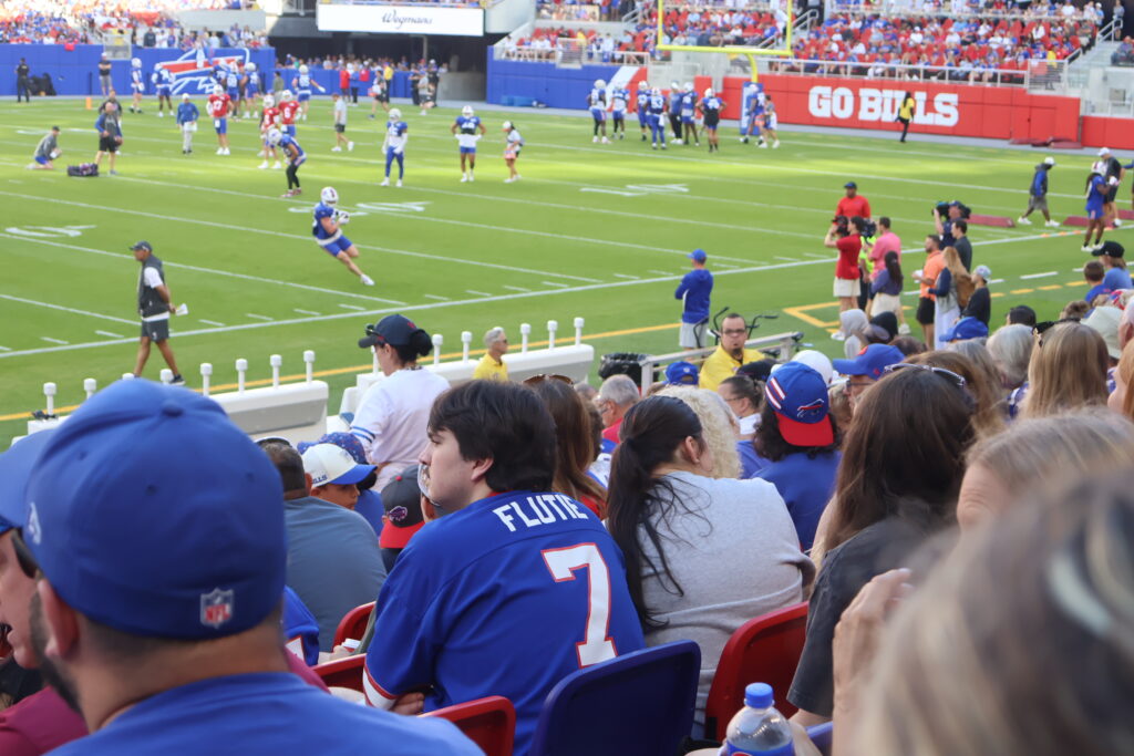 A fan in a Flutie number 7 jersey in the stands as players practice on the field