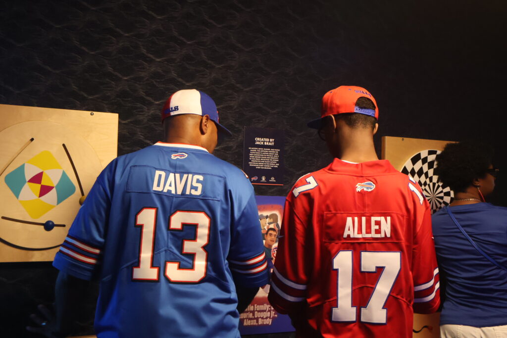 Two visitors in Buffalo Bills jerseys looking at a tactile activity wall in the sensory room