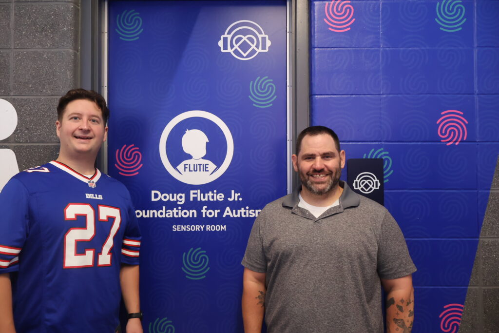 Two men standing beside the Doug Flutie Jr. Foundation for Autism sensory room banner