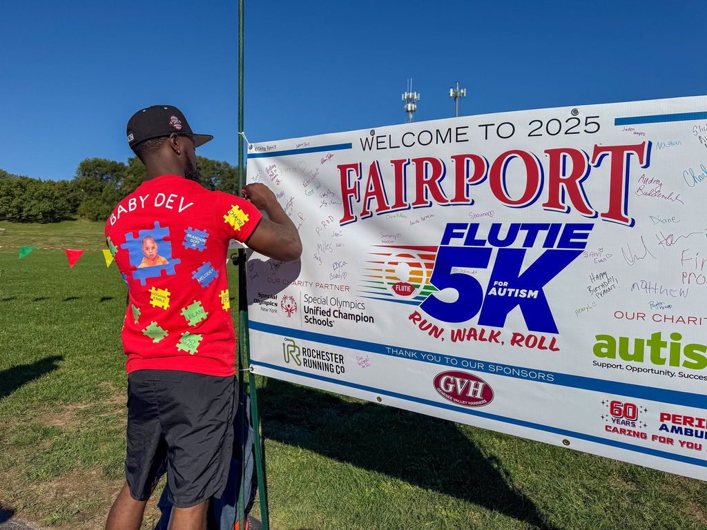 A runner signing the Fairport Flutie 5K welcome banner beside its sponsor panel