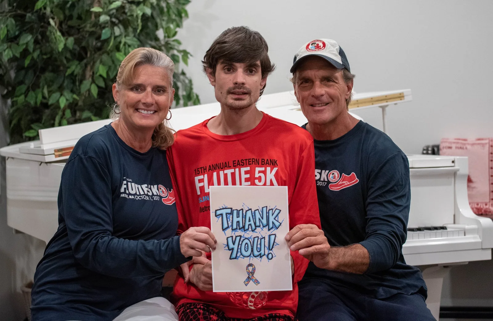 A family in Flutie 5K shirts sitting together, holding a hand-drawn 'Thank You!' sign with an autism awareness ribbon